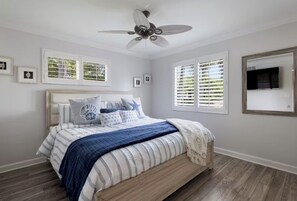 Ein Schlafzimmer mit Bett, Deckenventilator, Fenstern mit Jalousien und gerahmten Bildern an den Wänden.