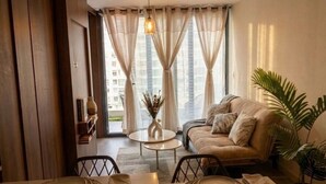 Una sala de estar con sofá, una mesa de comedor dispuesta con platos, un jarrón con flores secas y vistas a edificios a través de amplias ventanas.
