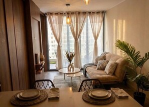 Ein Wohnzimmer mit einer Couch, einem Esstisch gedeckt mit Geschirr, einer Vase mit getrockneten Blumen und Blick auf Gebäude durch große Fenster.