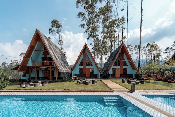 A poolside view of a resort with three triangular-roofed cabins, a clear blue pool, and a grassy area with seating.