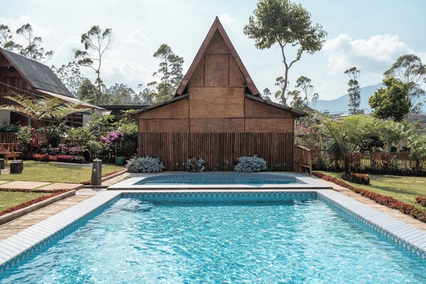 A swimming pool with a wooden deck, a triangular roof structure, and surrounding greenery.