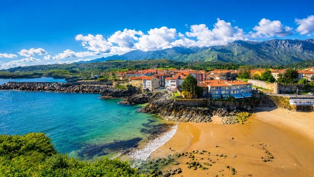 Una cittadina costiera con edifici lungo la spiaggia, un mare blu limpido e montagne sullo sfondo.