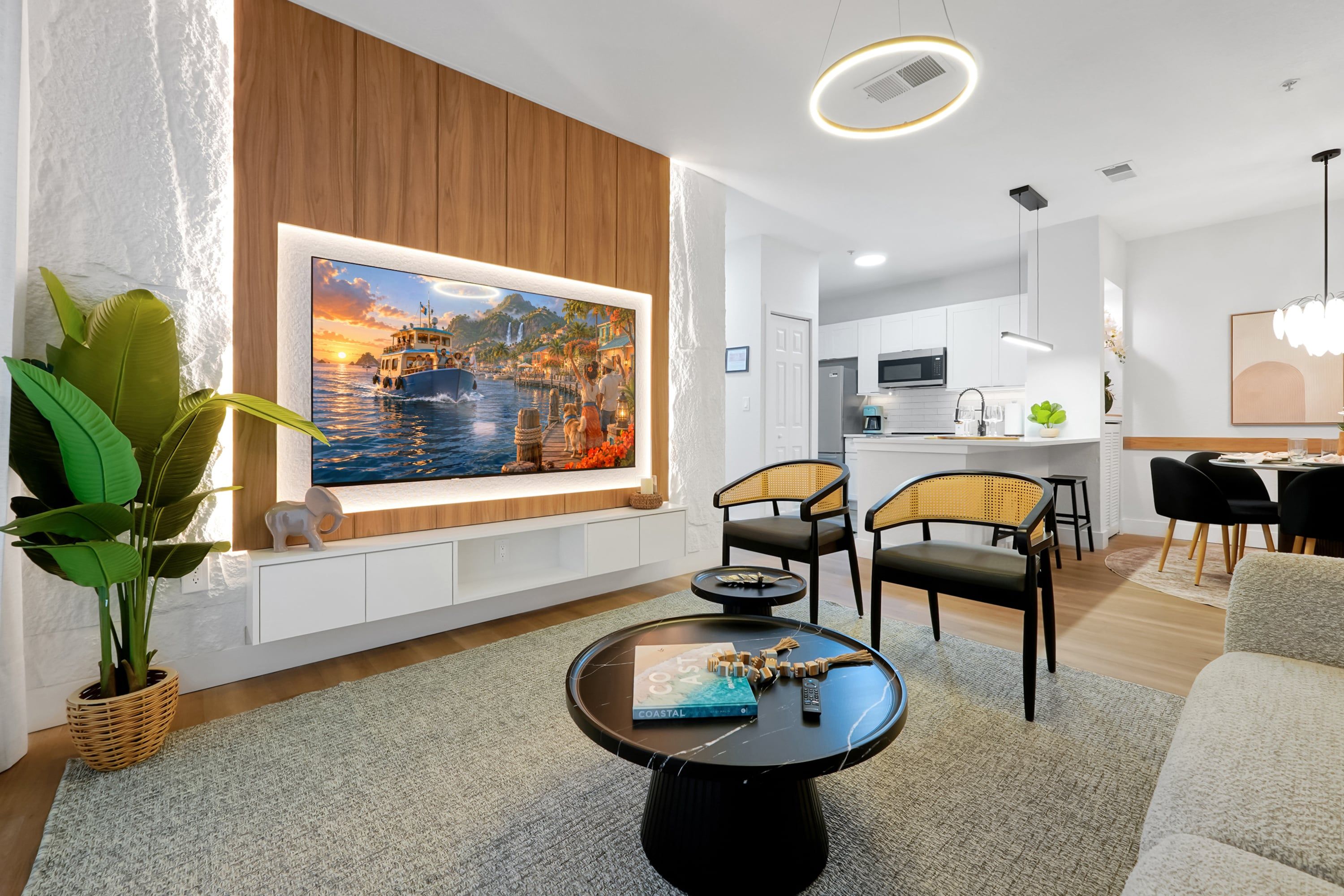 A modern living room with a large flat-screen TV, a dining area with a round table, and a kitchen in the background.