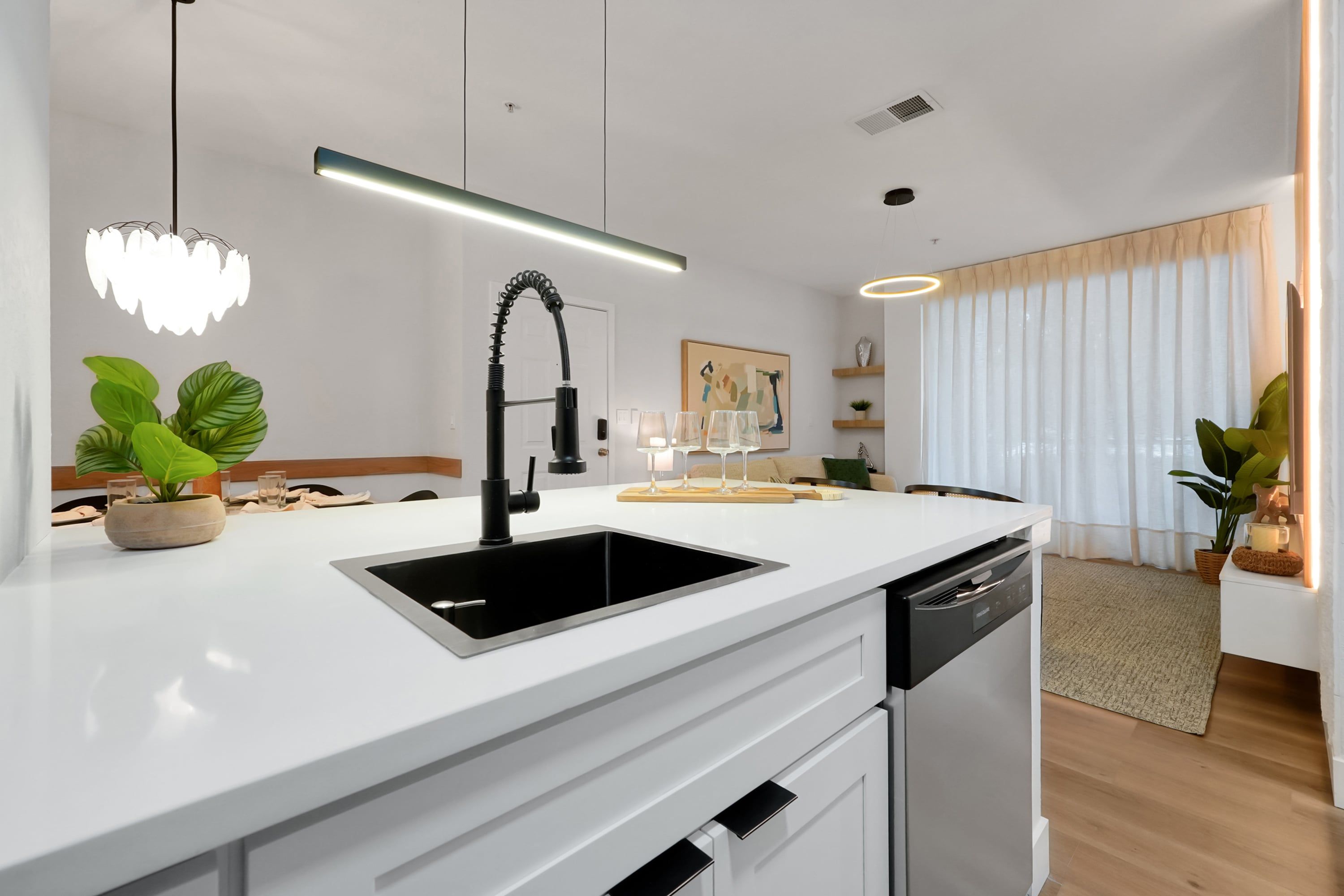 Eine moderne Küche mit weißer Arbeitsplatte, einem schwarzen Spülbecken und Edelstahlgeräten. Im Hintergrund sind Blumentöpfe und ein Essbereich zu sehen.