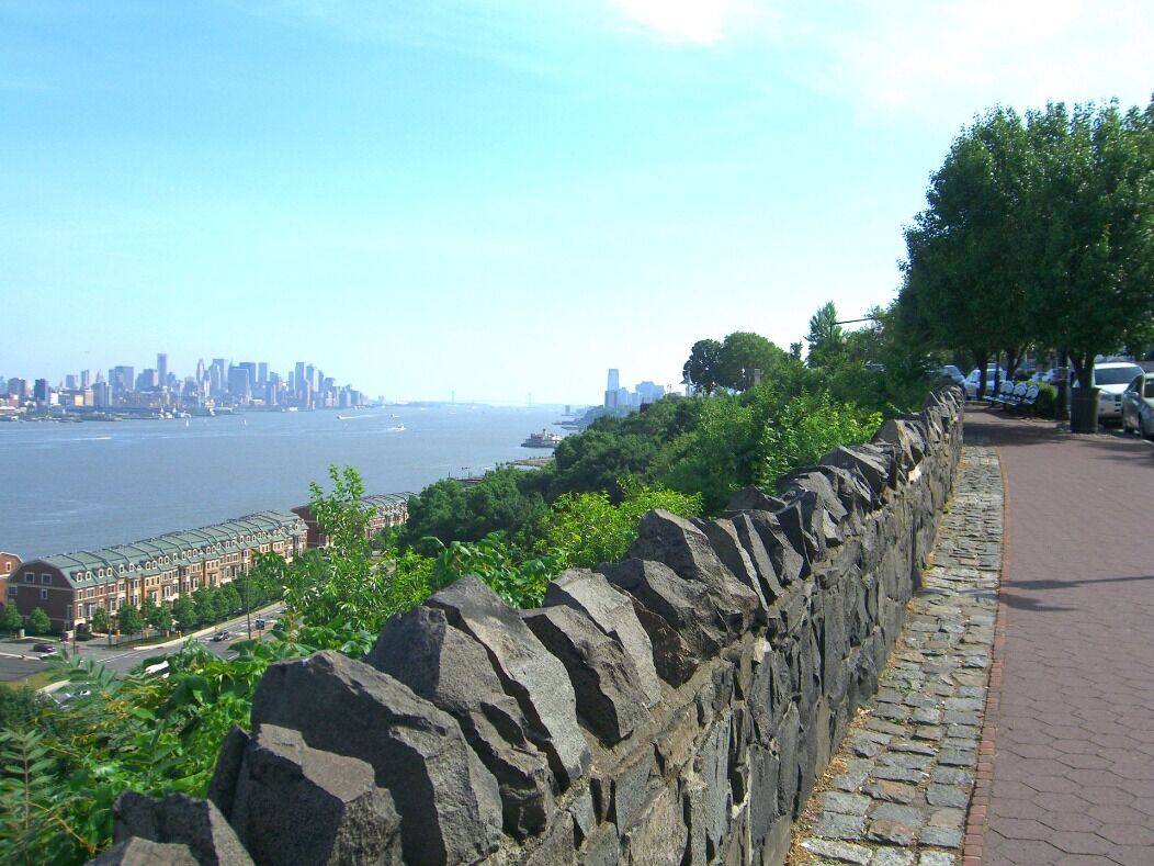 A stone wall with a paved walkway, overlooking a body of water and a distant city skyline.