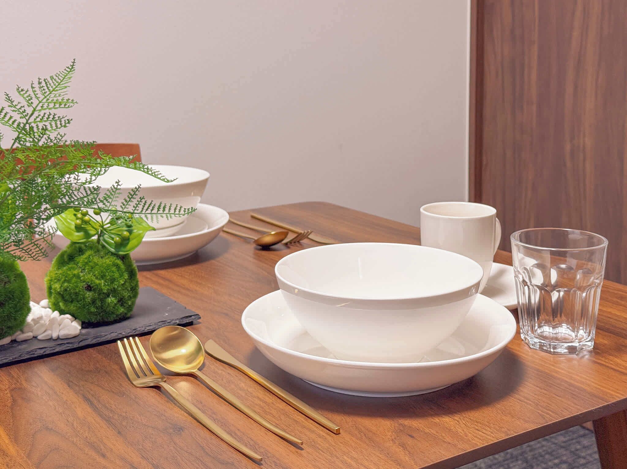 A neatly arranged dining table with white plates, bowls, a glass, and gold cutlery.