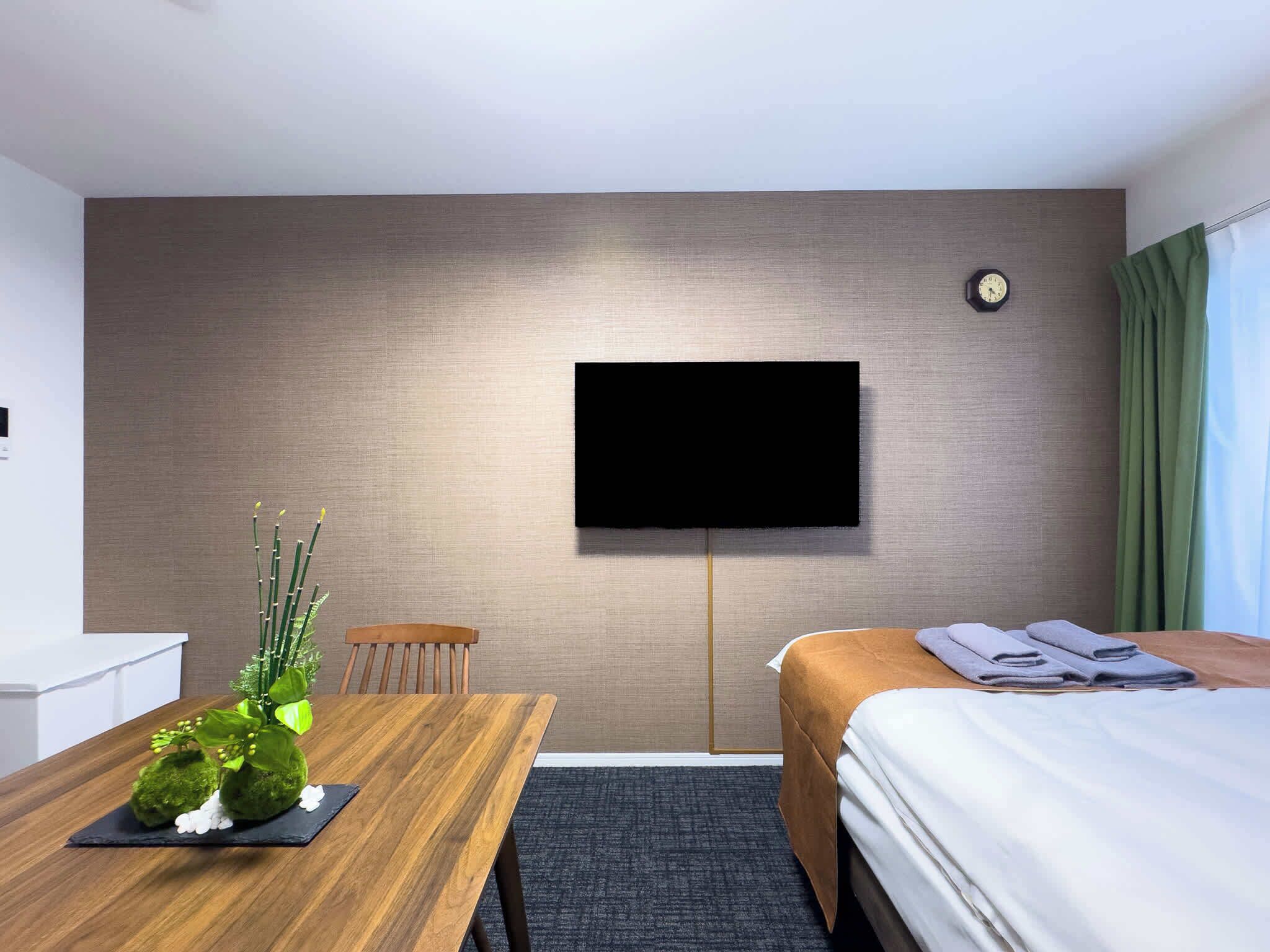 Habitación de hotel con cama, televisor montado en la pared, mesa de madera con una planta y una silla.