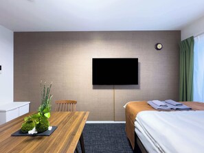 Une chambre d’hôtel avec un lit, un téléviseur fixé au mur, une table en bois ornée d’une plante et une chaise.