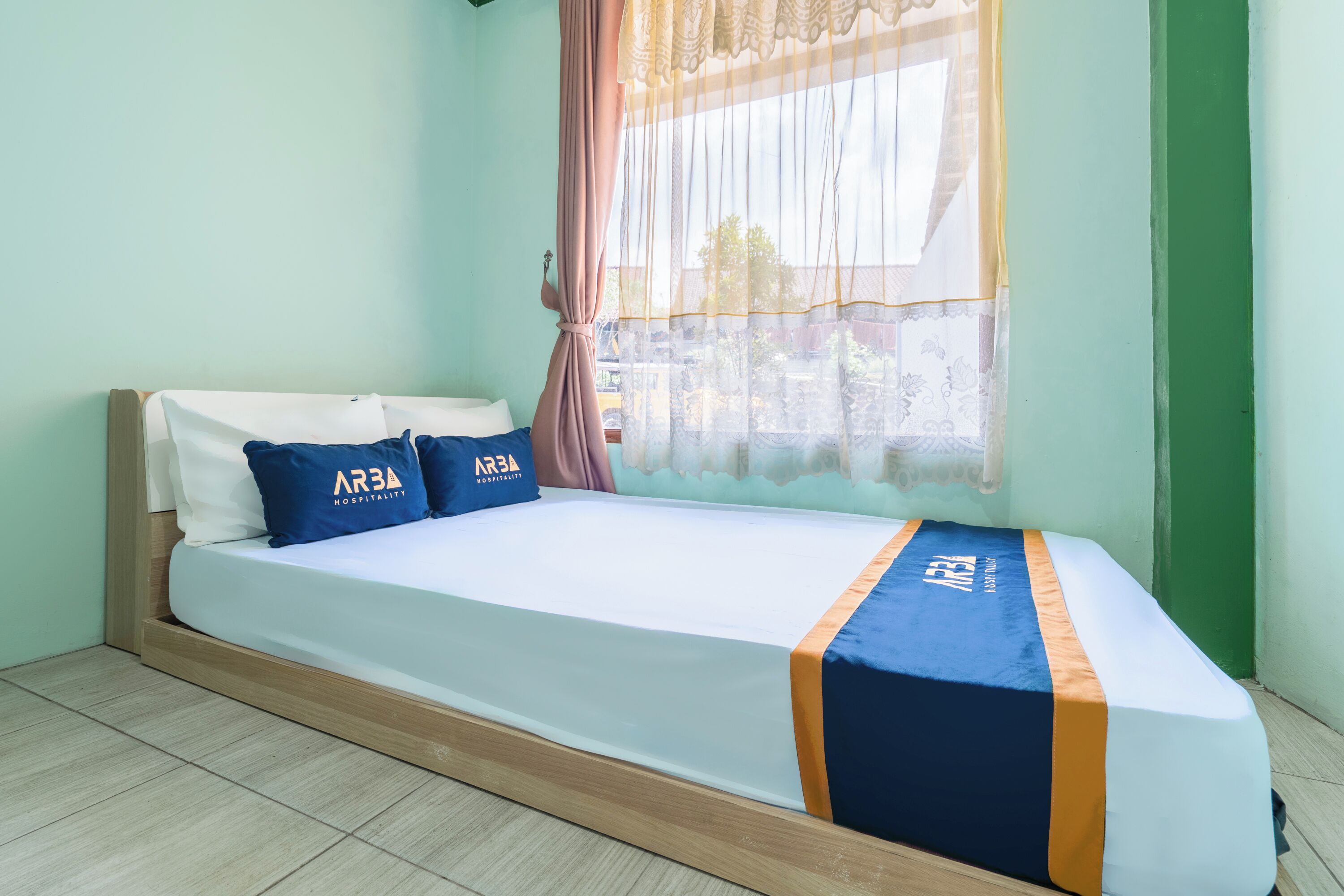 Una cama individual con colcha azul y naranja, dos almohadas con el logotipo de 'ARBA Hospitality', una ventana con cortinas traslúcidas y una pared verde.