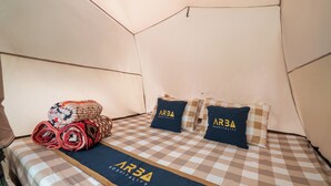 Ein Zeltraum mit kariertem Bettbezug, zusammengerollten Decken und Kissen mit dem Schriftzug 'AR3A Hospitality'.