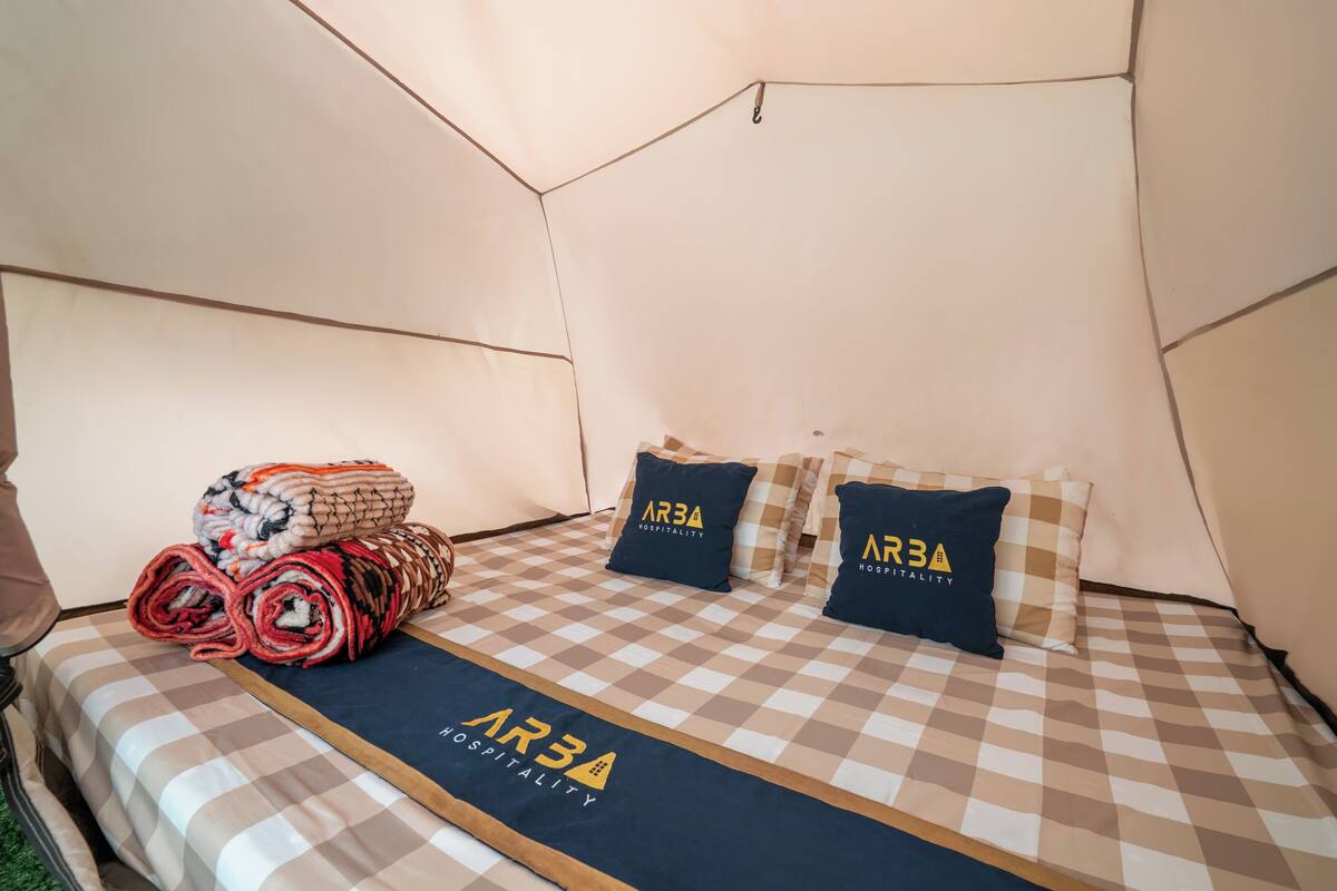 A tent-style room with a checkered bedspread, rolled blankets, and pillows with 'AR3A Hospitality' branding.