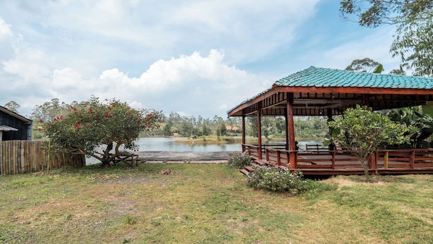 Ein hölzernes Gartenhaus mit grüner Dachdeckung, das auf einen See mit umgebenden Bäumen und einer Rasenfläche blickt.