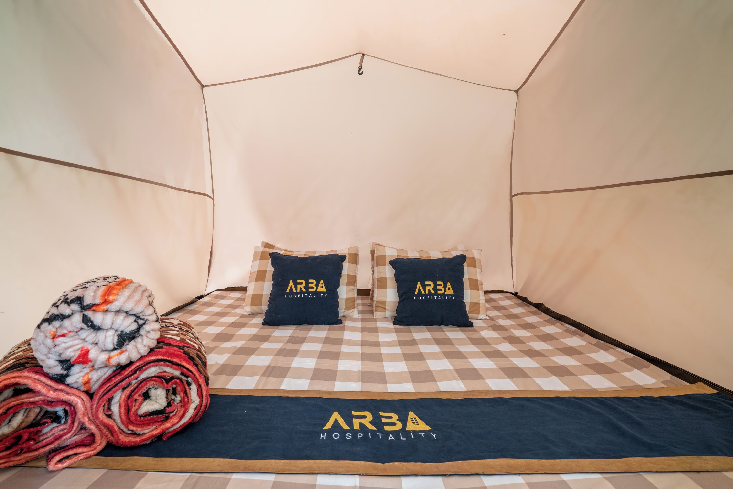 Een hotelkamer met een geruit sprei, twee kussens met het logo 'AR3A Hospitality', en een opgerolde plaid op het bed.