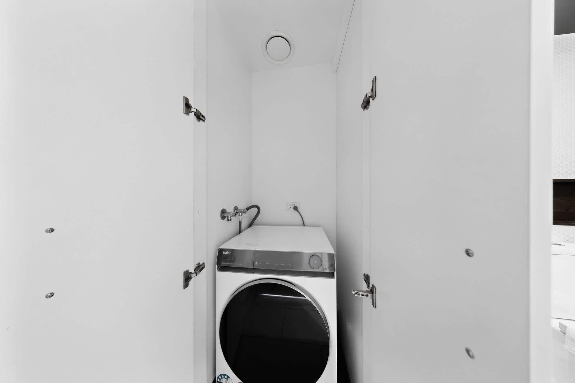 A compact, white laundry room with a front-loading washing machine, a small circular vent, and a power outlet on the wall.