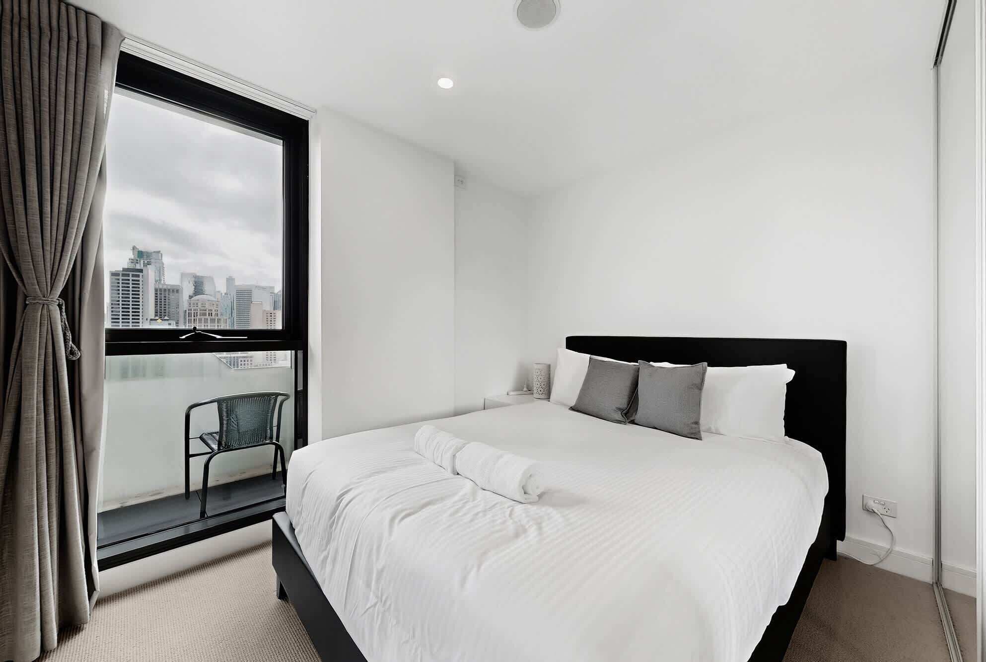 A hotel room with a large window, a bed with white bedding, a chair, and a city view.