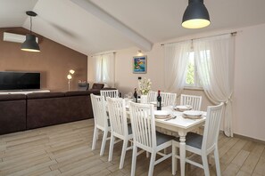Ein Essbereich mit einem weißen Tisch, gedeckt für vier Personen, weißen Stühlen, einem braunen Sofa und einem Fernseher im Hintergrund.