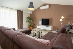 Ein modernes Wohnzimmer mit einer Sofagarnitur, einem Flachbildfernseher, einer Topfpflanze und einer Pendelleuchte.