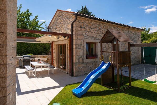 Ein Steinhaus mit Terrasse, Grill, Rutsche und Schaukel.