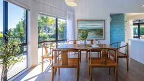 Uma área de refeições com mesa e cadeiras de madeira, janelas amplas, luminária pendente e um quadro na parede.