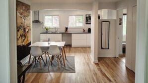 Een moderne keuken met witte kastjes, een eettafel met stoelen, een groot raam en een schilderij aan de muur.