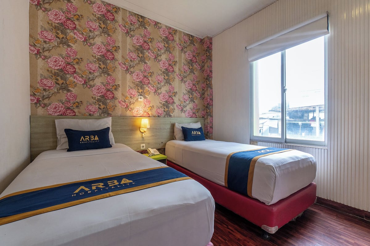 Une chambre d’hôtel avec deux lits, un papier peint à motifs floraux, une fenêtre donnant sur un beau paysage et un logo imprimé sur les draps.