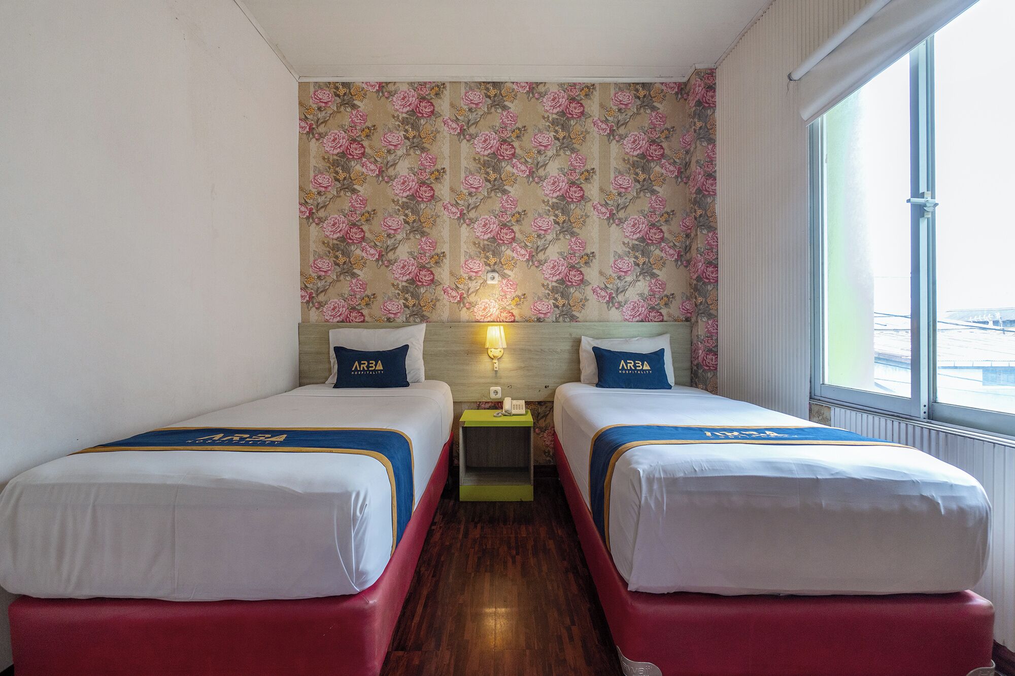 A hotel room with two beds, floral wallpaper, a window with a view, and a small table with a lamp.