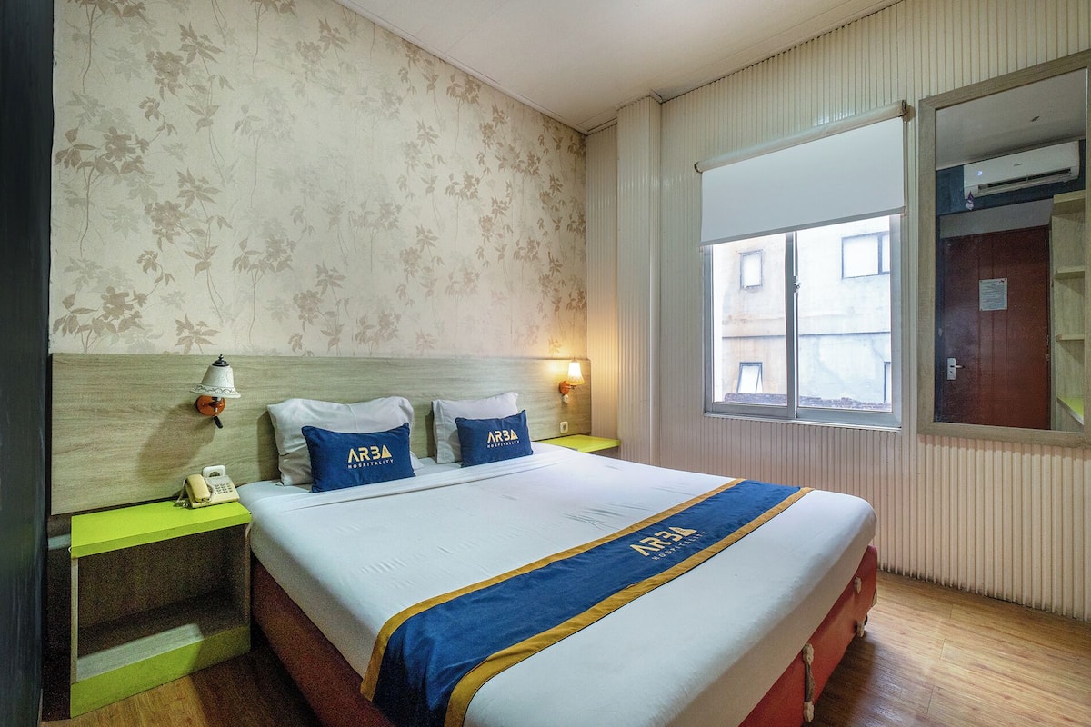 Une chambre d’hôtel avec un lit, deux oreillers ornés de logos, une lampe de chevet, une fenêtre et un mur recouvert d’un papier peint à motifs floraux.