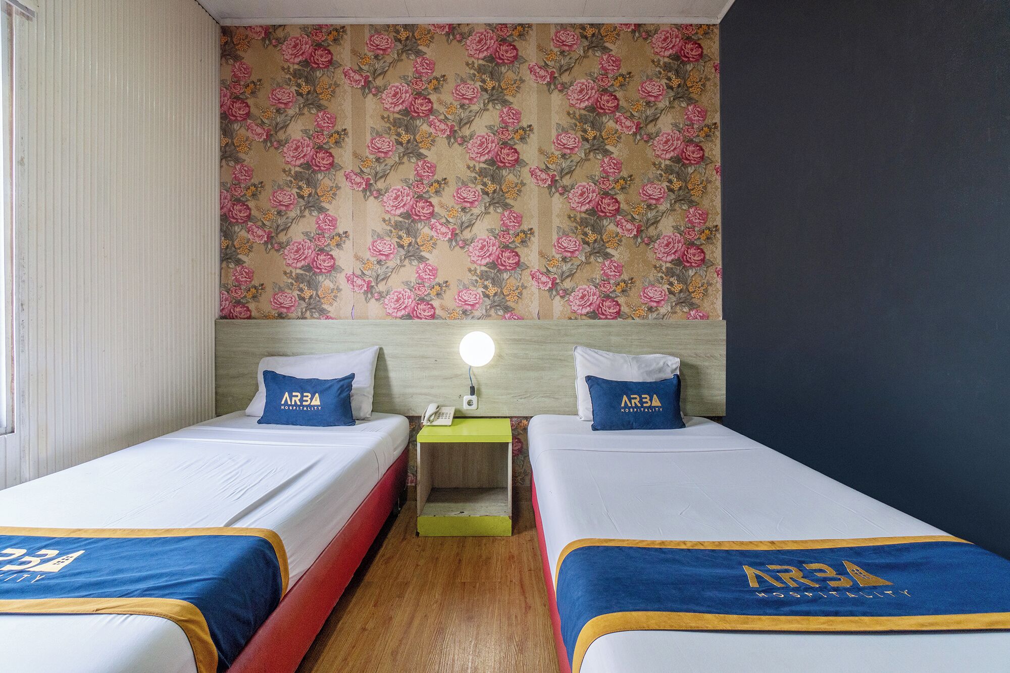 A hotel room with two beds, floral wallpaper, a nightstand with a lamp, and a wooden headboard.
