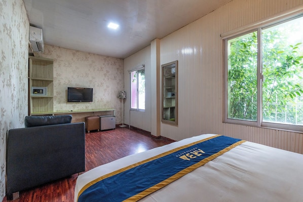 A hotel room with a bed, a desk with a television, a chair, and a window with a view of trees.