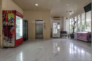 A clean lobby with a Coca-Cola vending machine, an elevator, a vending machine with snacks, a bench, and a potted plant.