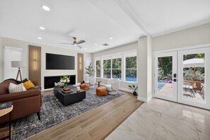 A modern living room with a fireplace, a flat-screen TV, and a large window overlooking a pool. - The Woodlands Luxe 5BR/4.5BA Heated Pool and Grill (Spring)