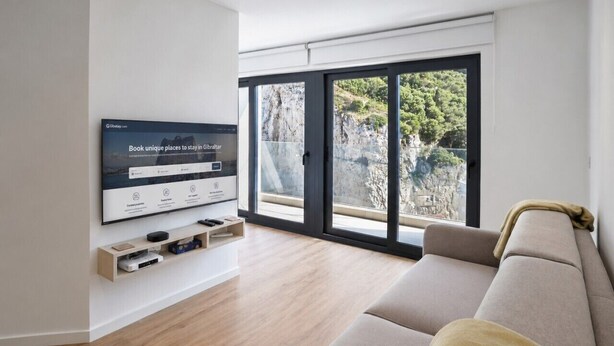A modern living room with a large TV, wooden flooring, a beige sofa, and a view through large windows.