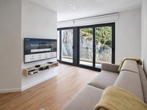 A modern living room with a large TV, wooden flooring, a beige sofa, and a view through large windows.
