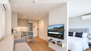 A modern hotel room with a sofa, a bed, a TV, a desk, and a kitchen area.