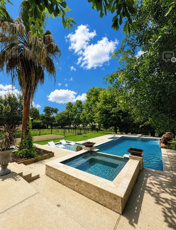 Una piscina rectangular con un jacuzzi, rodeada por un solárium de piedra y un césped cuidado.