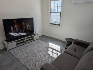 A living room with a flat-screen TV, a grey sofa, a small side table, and a window with a view.