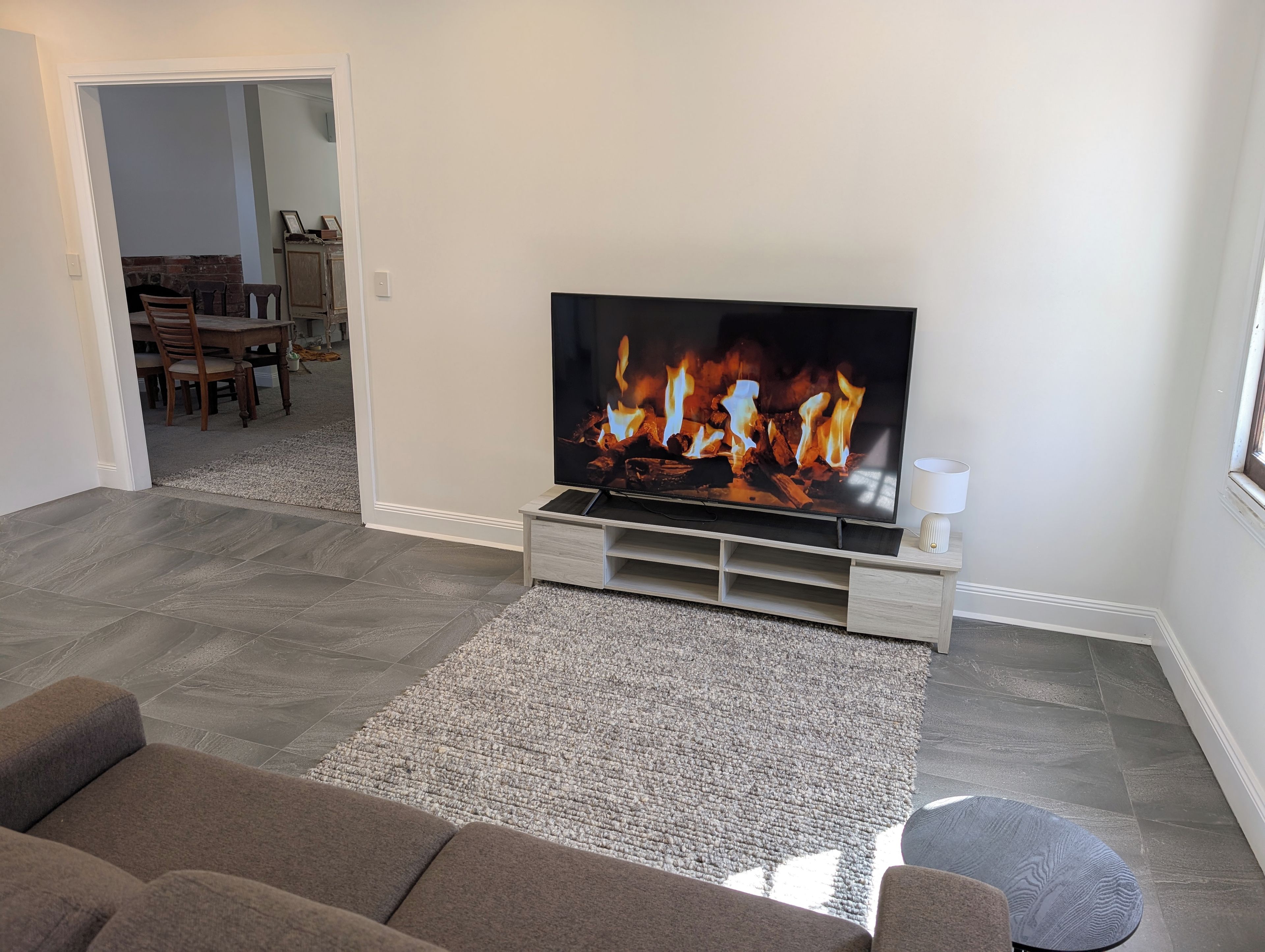 Een woonkamer met een flatscreen-televisie waarop een haard wordt getoond, een grijs tapijt met patroon en een bruine bank.