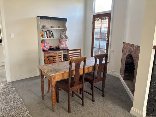 A dining area with a wooden table and chairs, a bookshelf with books and a stuffed toy, and a brick fireplace.