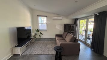 Una sala moderna con un televisor de pantalla plana, un sofá marrón, una mesita auxiliar pequeña, una planta en maceta y un puerta corrediza de vidrio que da a un área exterior.