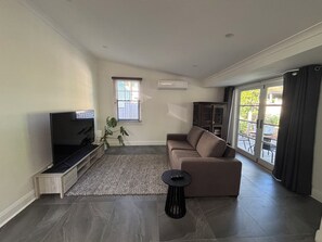 A modern living room with a flat-screen TV, a brown sofa, a small side table, a potted plant, and a sliding glass door leading to an outdoor area.