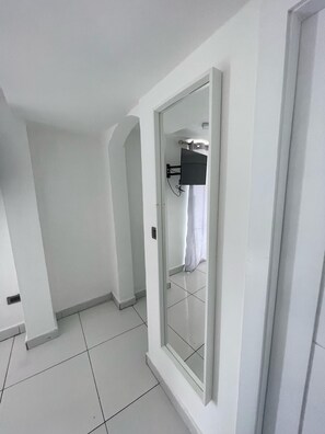 A hallway with a white tiled floor, a door with a mirror, and a mounted television. - Modern 2BR Penthouse with Jacuzzi & City View (Santo Domingo)