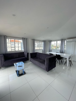 A modern living room with a sofa, a coffee table, and a dining area with chairs and a table. - Modern 2BR Penthouse with Jacuzzi & City View (Santo Domingo)