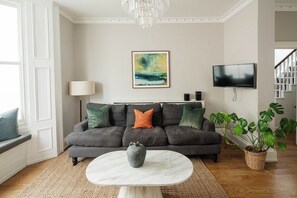 Ein Wohnzimmer mit grauem Sofa, weißem Couchtisch, einem Gemälde, einem Fernseher und einer Treppe.