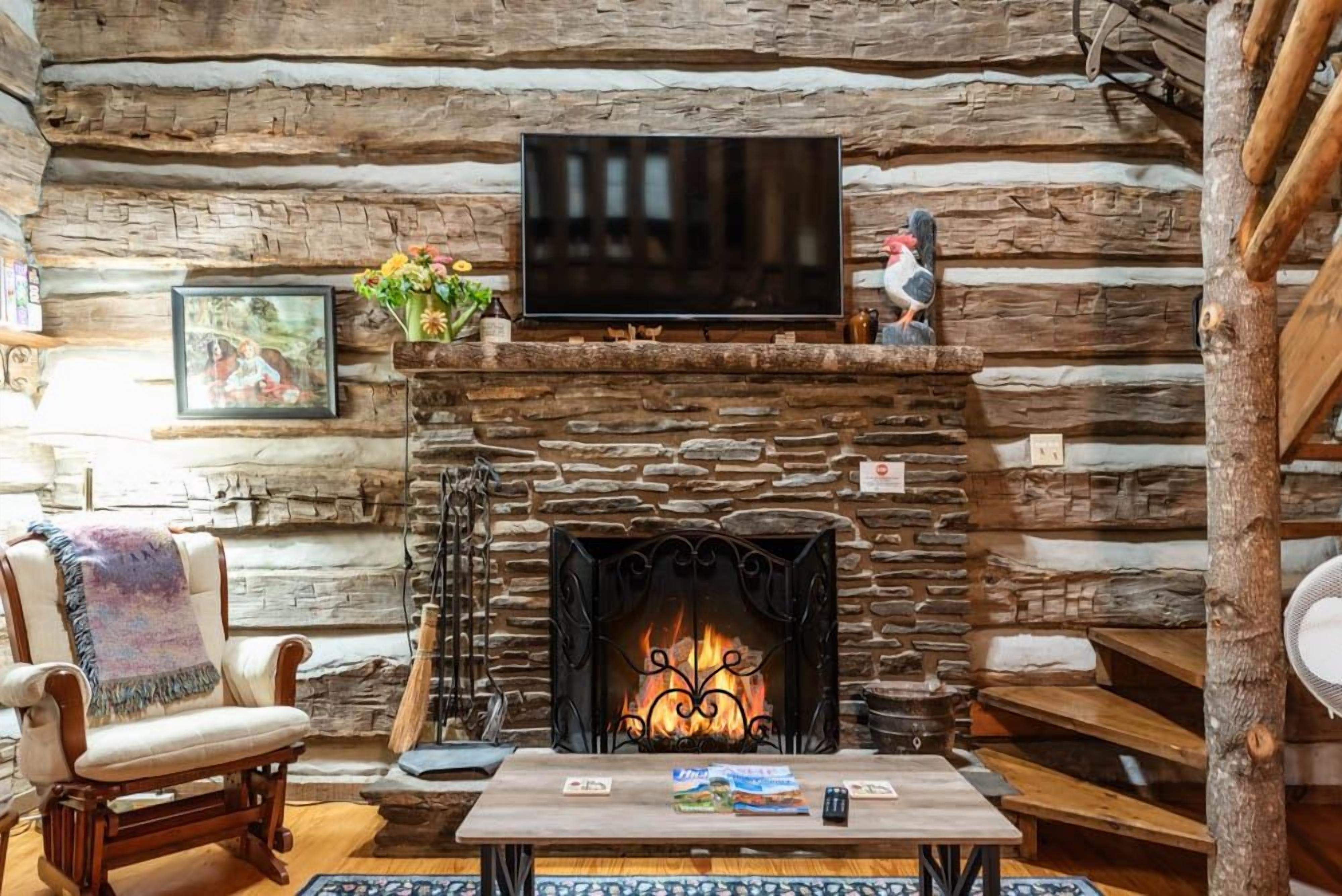 A log cabin interior with a stone fireplace, a television mounted above it, and a cozy seating area with a chair and a table.