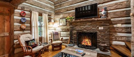Interior de una cabaña de troncos con chimenea de piedra, muebles de madera y un televisor de pantalla plana.