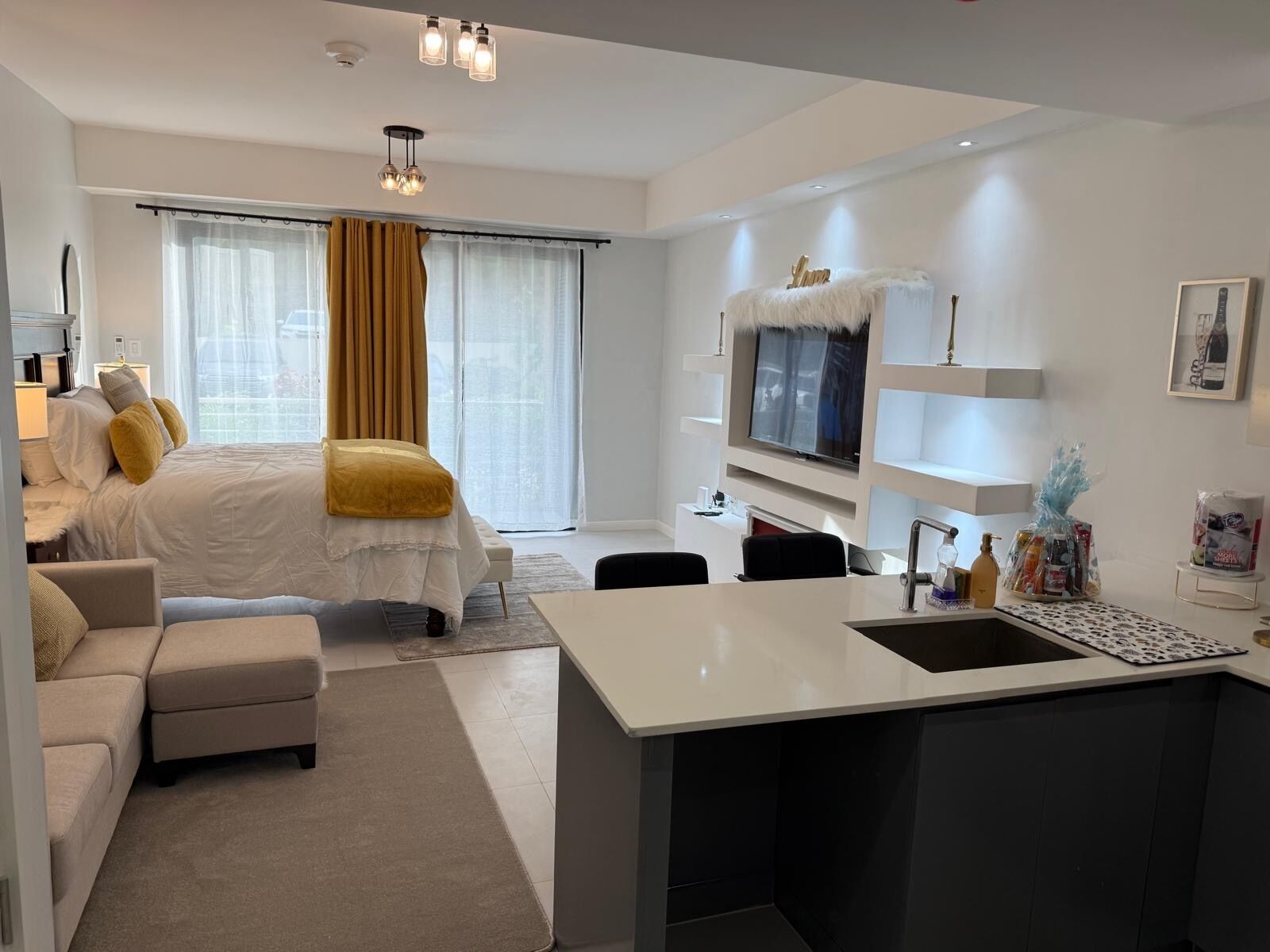 A modern hotel room with a bed, a sofa, a small kitchen area, and a TV.