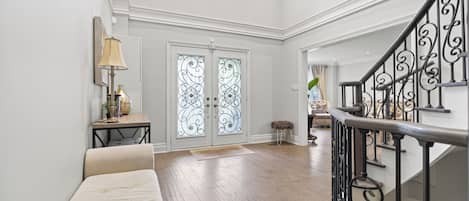 A spacious foyer with a high ceiling, a staircase with ornate metal railings, and a pair of double doors with decorative glass panels.