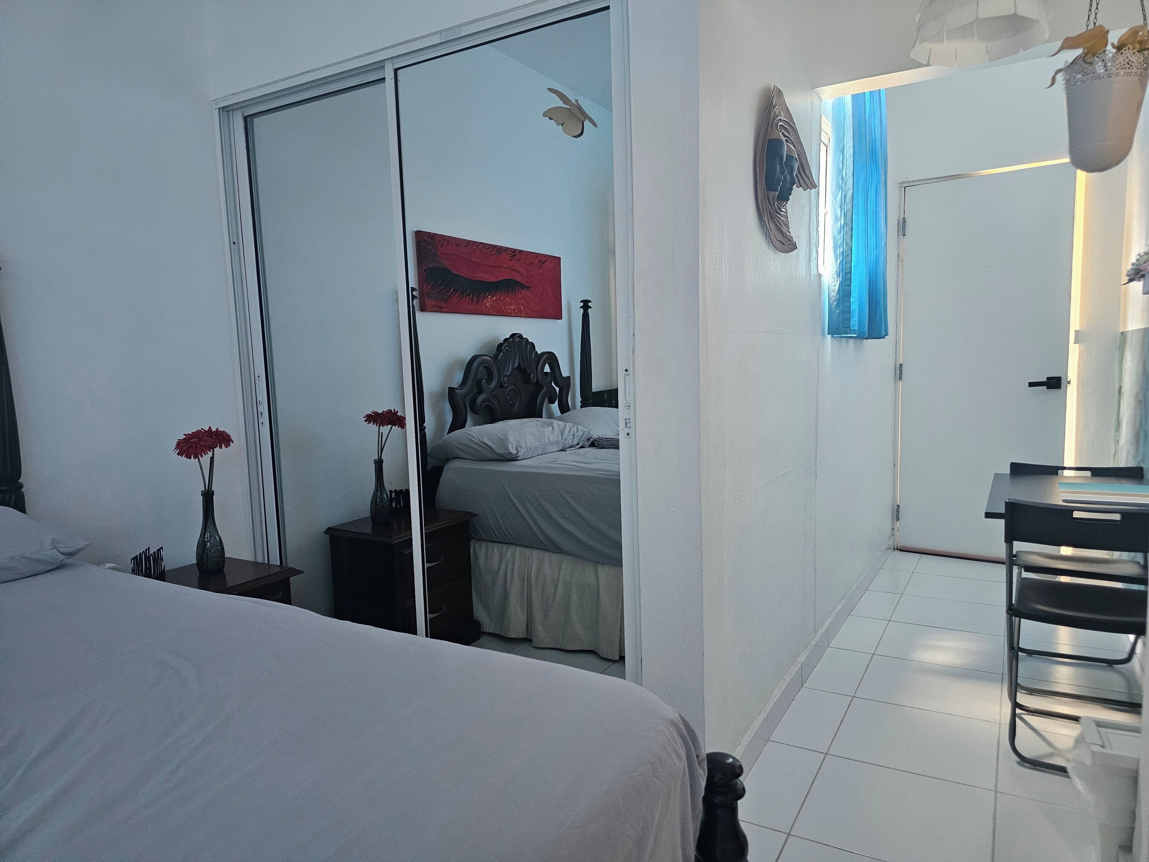 Une chambre à coucher avec un lit, une table de chevet ornée d’un vase, un miroir, une porte donnant sur une autre pièce et une œuvre d’art accrochée au mur.
