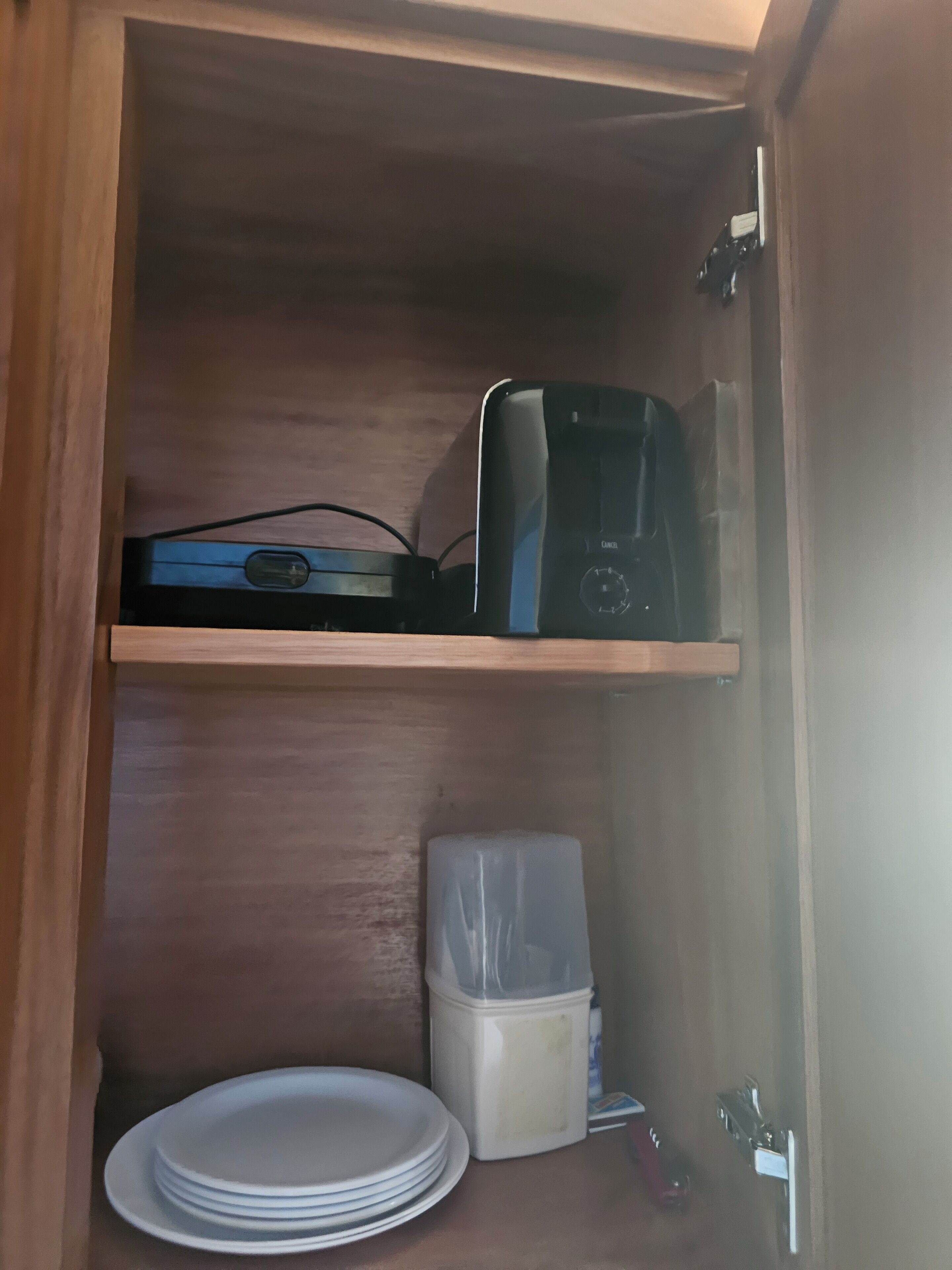 Un meuble en bois sur lequel se trouve, sur une étagère, un appareil électronique noir, ainsi qu’une pile d’assiettes blanches et un récipient transparent en plastique.