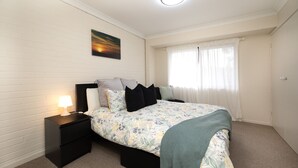 A bedroom with a bed, nightstands, a lamp, a painting, and a window with curtains. - The Nook in Mollymook - Walk to the beach! (Mollymook Beach)
